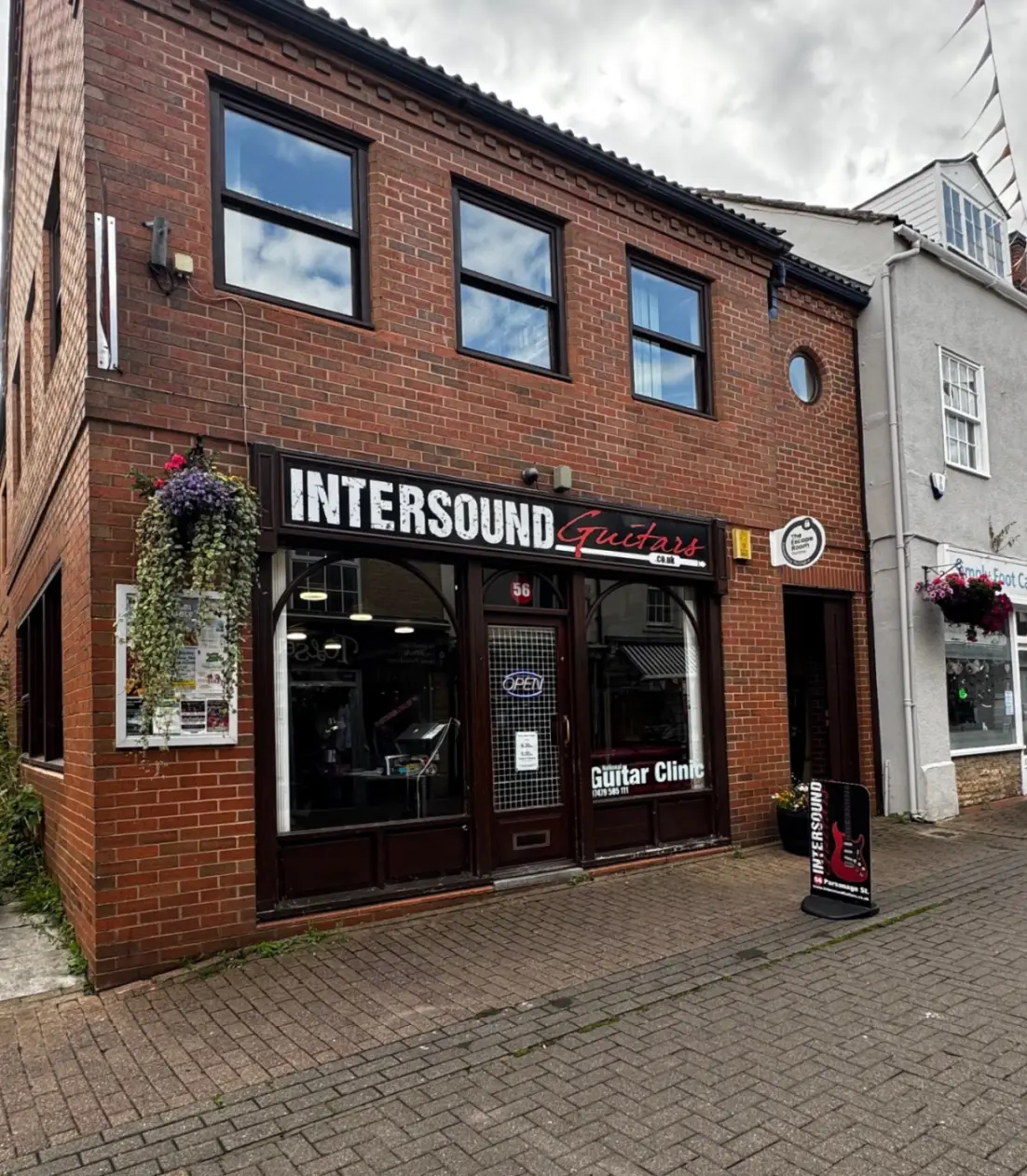 Front of red bricked shop on the high street called Intersound Guitars with signage and floral hanging basket on the wall.